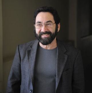 Man standing from waist up dressed all in black with black hair and beard plus glasses.