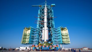 A tall white rocket with two little side boosters stands next to a wide blue assembly scafolding, swung open to allow the rocket to move.