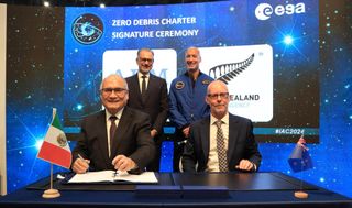 Four men behind a black table, two sitting, two standing behind them. two small flags are at the foreground corners of the table. A backdrop of space stands behind them.