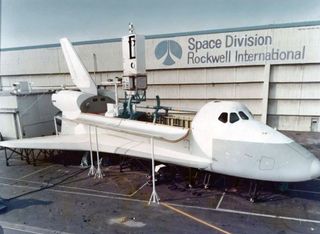 side view of a white space shuttle mockup, in front of a wall emblazoned with the words" space division, rockwell international"