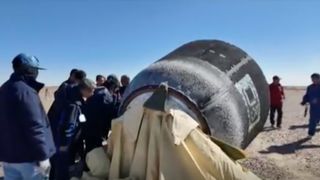 a group of people inspect a gray cylindrical spacecraft in a desert landscape