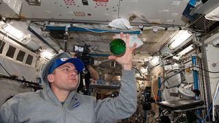 an astronaut in a hoodie stretching out his hand around a green sphere of water floating in space