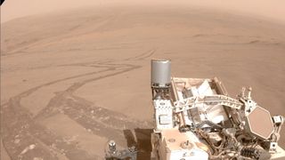 photo of a mars rover in the foreground, with its tracks in the red dirt stretching off into the distance