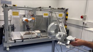 photo of a hand operating, at a distance, a dirt-scooping robot inside a laboratory