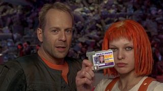 a woman with medium-length red hair and a man with stubble and light, thin hair look at the camera while the woman holds an ID card with her face on it