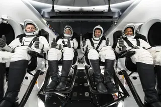 four astronauts, three men and one woman, giving thumbs up in black and white spacesuits