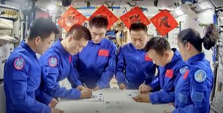 six astronauts in blue jumpsuits crowd around a table in a bright spacecraft. two across from one another each sign documents.