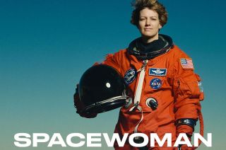 a woman in an orange flight suit stands outside, with the word "spacewoman" in white across the bottom of the photo