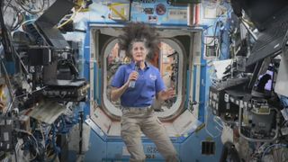 a woman in a blue shirt and khaki pants holds a microphone aboard the international space station