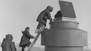 People climb a ladder resting on the side of a pipe, standing burried in the snow.