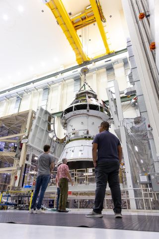 a spacecraft on a large manufacturing scaffolding surrounded by people who are looking at it, backs to the camera