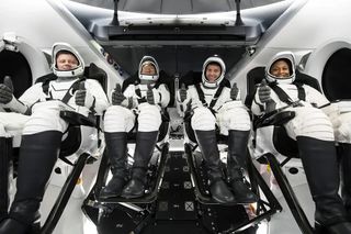 four people in black and white spacesuits sit inside a space capsule