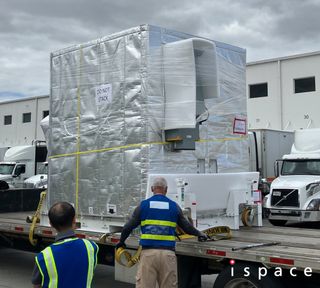 Two balding men in highlighter vests attend a large metal box secured to the flatbed trailer of a semi.