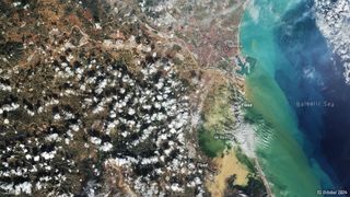 Aerial view from a high altitude, with green populated landmass on the left, and the Balearic Sea on the right. Some small white clouds are sparingly scattered