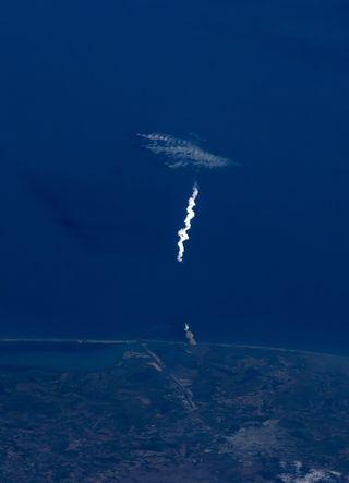 A white squiggly cloud scrapes vertically in the center, with the dark blue of the sea dropped behind. Land cuts across the bottom, its green hue blued by the atmosphere above.