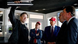 a man holds up a small model of a spacecraft while four men in suits look on