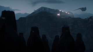 Many robed and veiled women standing in front of a cliff face, with a spaceship flying overhead and a building atop the cliff.