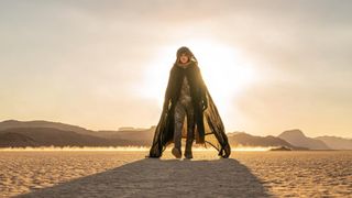 A man with blue eyes and a hood is wearing a long flowing garment and a clip reaching from his nose past the side of his face while walking on sand with the sun in the background.