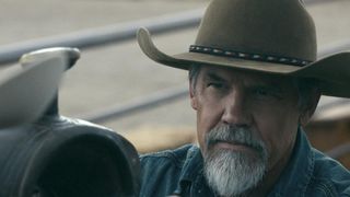 A man with a grey goatee bears stands next to a saddle while wearing a beige cowboy hat.