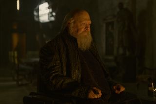 A grey-bearded old man in a dark stone room