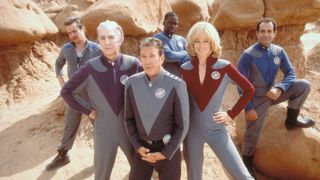 Four men, a woman, and an alien man wearing sci-fi uniforms stood in a rocky desert area.