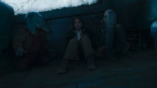 Still from Star Wars: Skeleton Crew TV series. Three children – Neel (blue elephant-like alien boy), Fern (human girl), and KB (girl with an asymmetrical white bob and wearing visor across her eyes) – are sitting with their backs against a wall in a dark room, hiding.