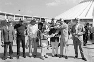 A Mickey Mouse costumed performer welcomes a group of men in jackets and slacks outside Space Mountain at Walt Disney World.